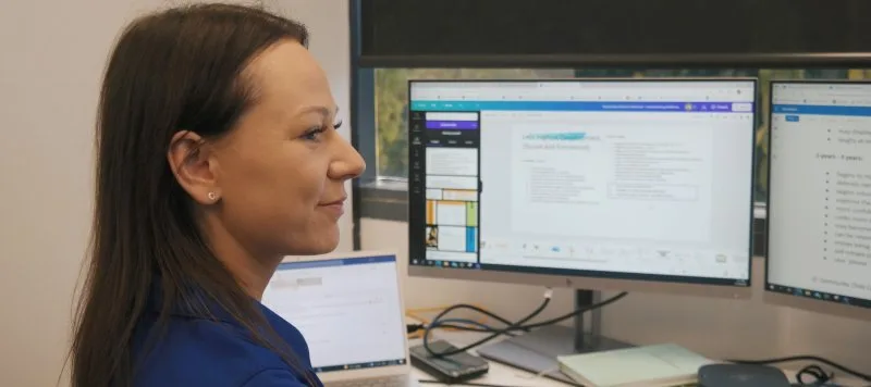 Staff member working on a document on a dual screen computer. She is smiling