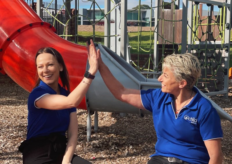 Two Child Australia staff members high fiving and smiling in front of an outdoor playground with a red slide, the sun is shining