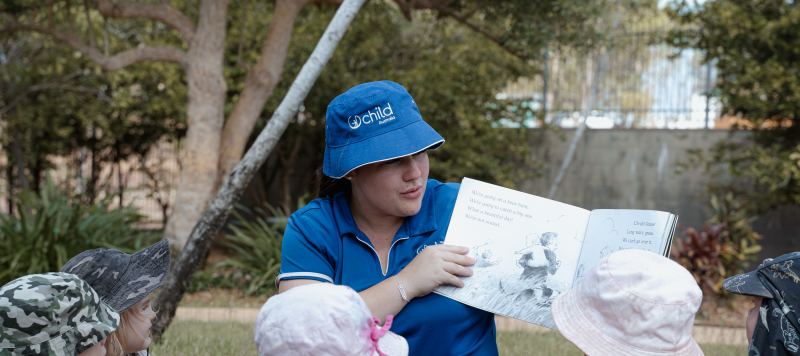 A teacher wearing a Child Australia hat reading and showing illustrations to a group of toddler age students outdoors during a Book Corner activity. The students are looking interested with the book while wearing hats