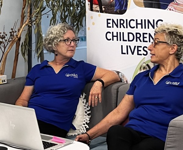 Two Child Australia staff members sitting on a coach having a conversation in front of a laptop. A sign behind them says 