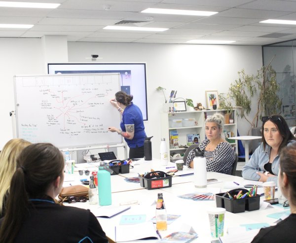 A group of women sit at a desk whilst one writes on a white board